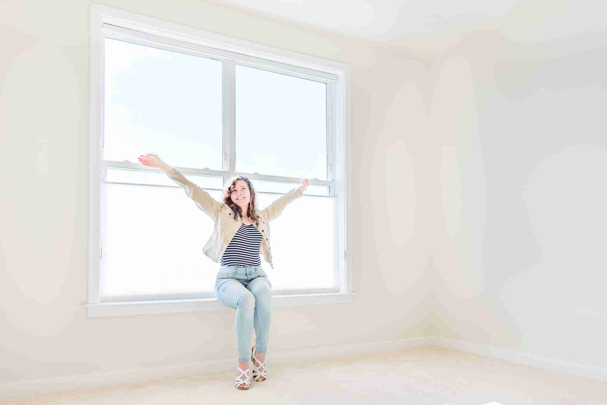 One young woman sitting on windowsill in white, bright empty, clean room on carpet of modern new house, home, apartment sml